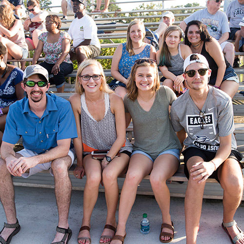 four students sitting on bleachers with many people behind them