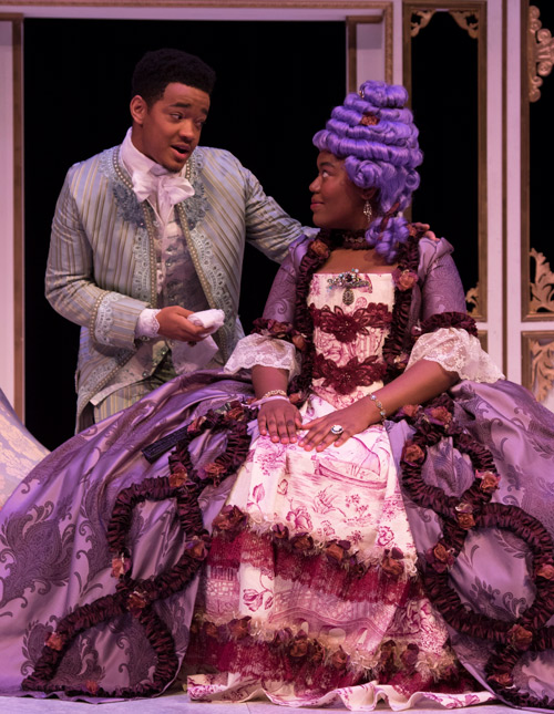 woman in large pompadour dress and bee hive hair sitting and talking with a man in similar period dress