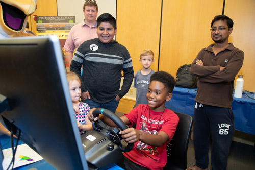 boy playing a driving game on a computer while a group of people watch on